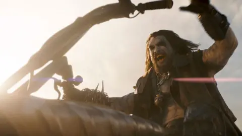 DC Studios A screengrab from Supergirl shows Jason Momoa as Lobo, dressed in a leather vest, riding a motorbike and roaring as he speeds towards his destination.