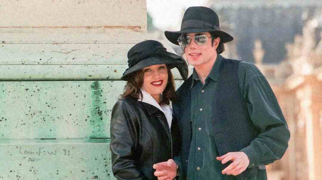 A man and woman stand side by side outdoors, both wearing black hats and dark clothing. The woman smiles while the man wears sunglasses and also smiles slightly. They are near a light-colored stone structure.