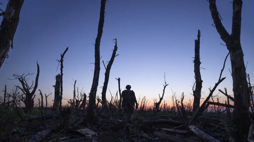 A soldier silhouetted against a landscape of burned trees as the sun sits at the horizon.