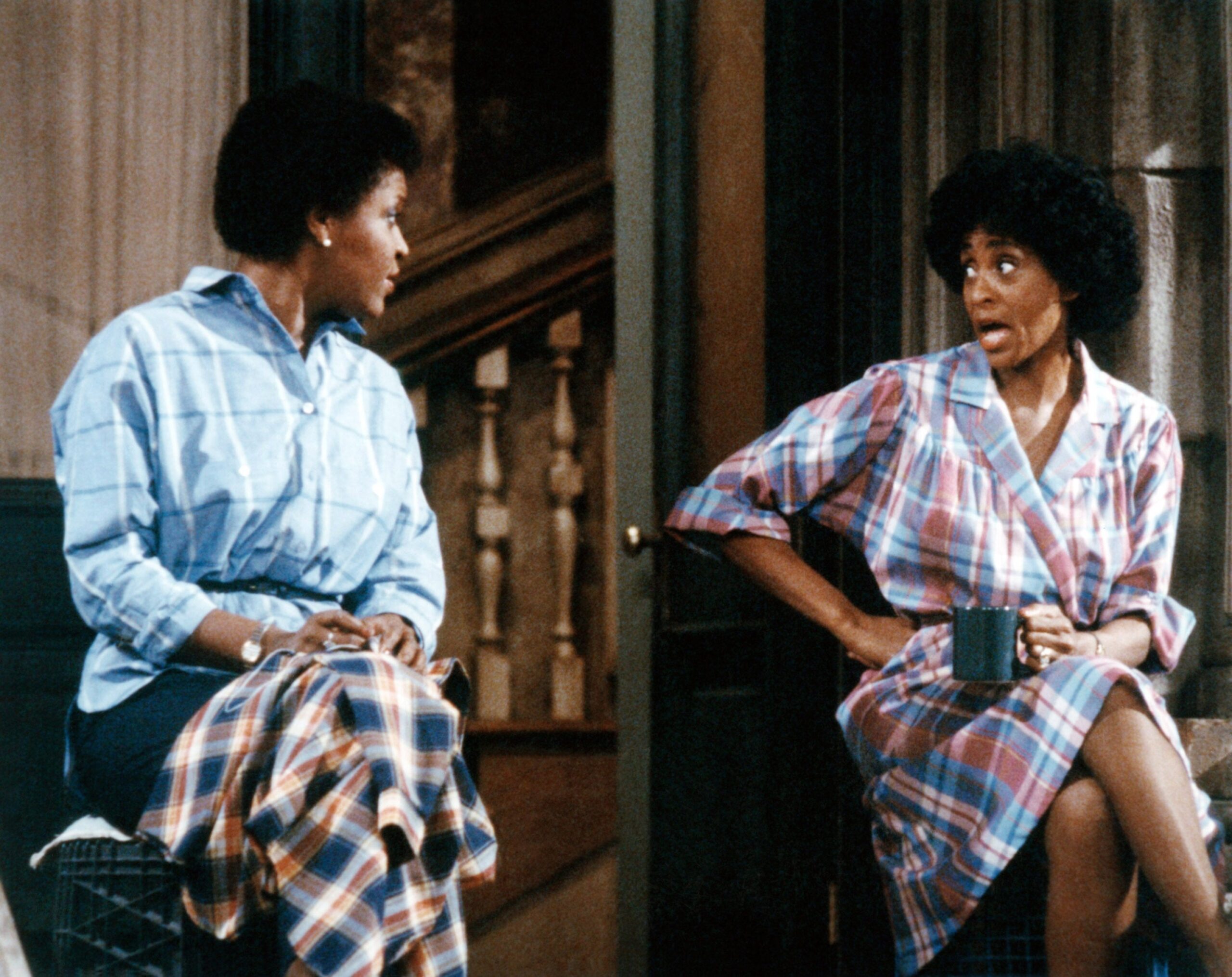 Two people sit on a porch, engaged in conversation. Both wear plaid clothing. One holds a mug, looking surprised. The setting suggests a candid moment
