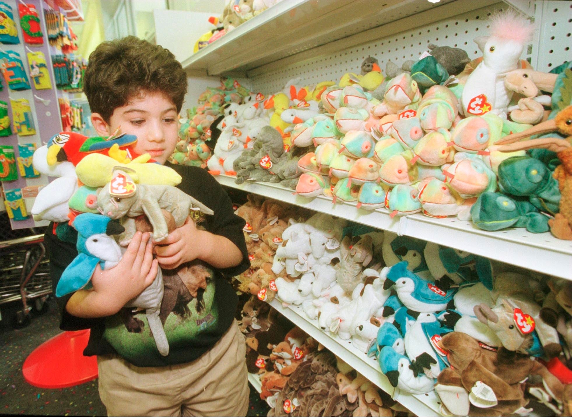 A young boy holds a stack of Beanie Babies while shopping at a toy store filled with shelves of the popular stuffed animals