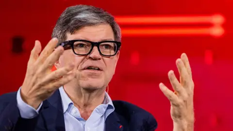 Bloomberg via Getty Images Yann LeCun wears fashionable thick-rimmed glasses, a light blue shirt and navy jacket. He is using his hands to articulate a point while speaking at a conference.