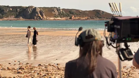 BBC A camera man is seen from behind filminf two women walking along a sandy beach with cliffs in the background.
