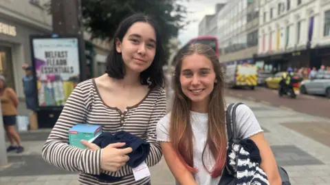 BBC Two teenage girls. the one on the left has a black bob, and a stripey top. The one on the right has long light brown hair, a whit t shirt and a shoulder bag,