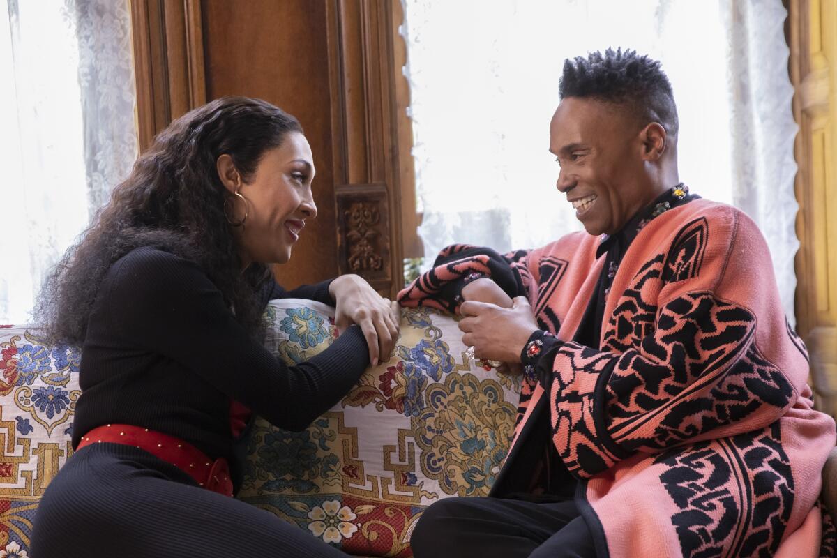 A woman in a black dress and red belt sits on a couch with a man in a pink and black sweater.