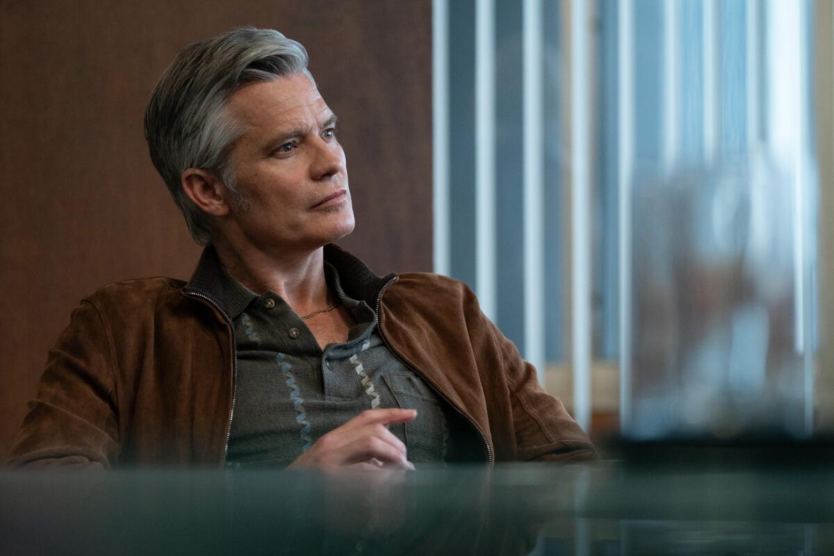 A man with salt and pepper hair and a brown jacket sits in a chair while looking off to his left