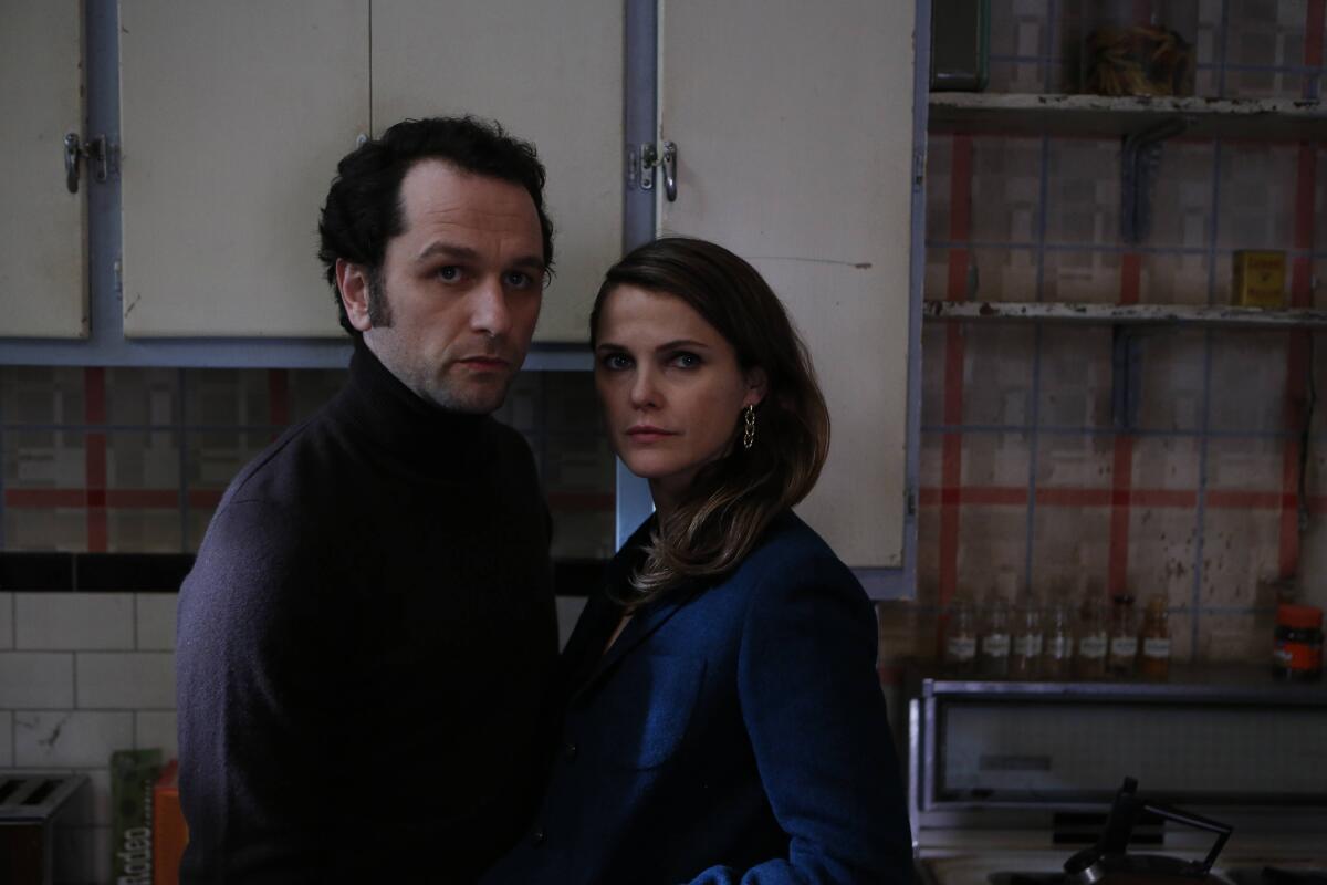 A man in purple shirt stands next to a woman in a blue top in a run-down kitchen.