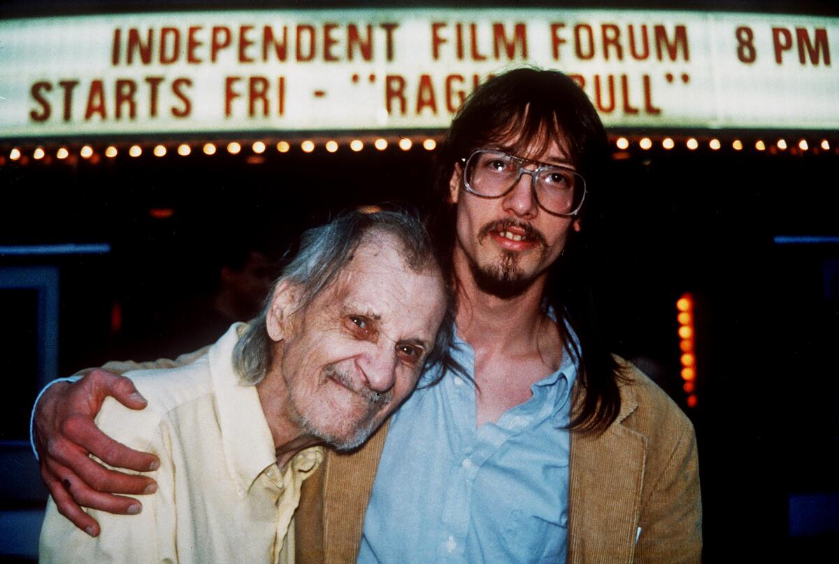 An older man in a yellow shirt leans on the shoulder of a younger man with long brown hair and glasses.