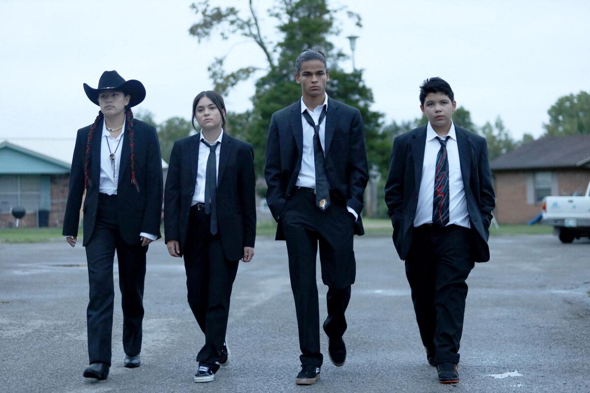 Four teenagers in black suits, ties and sneakers walk down the middle of the street of a neighborhood.