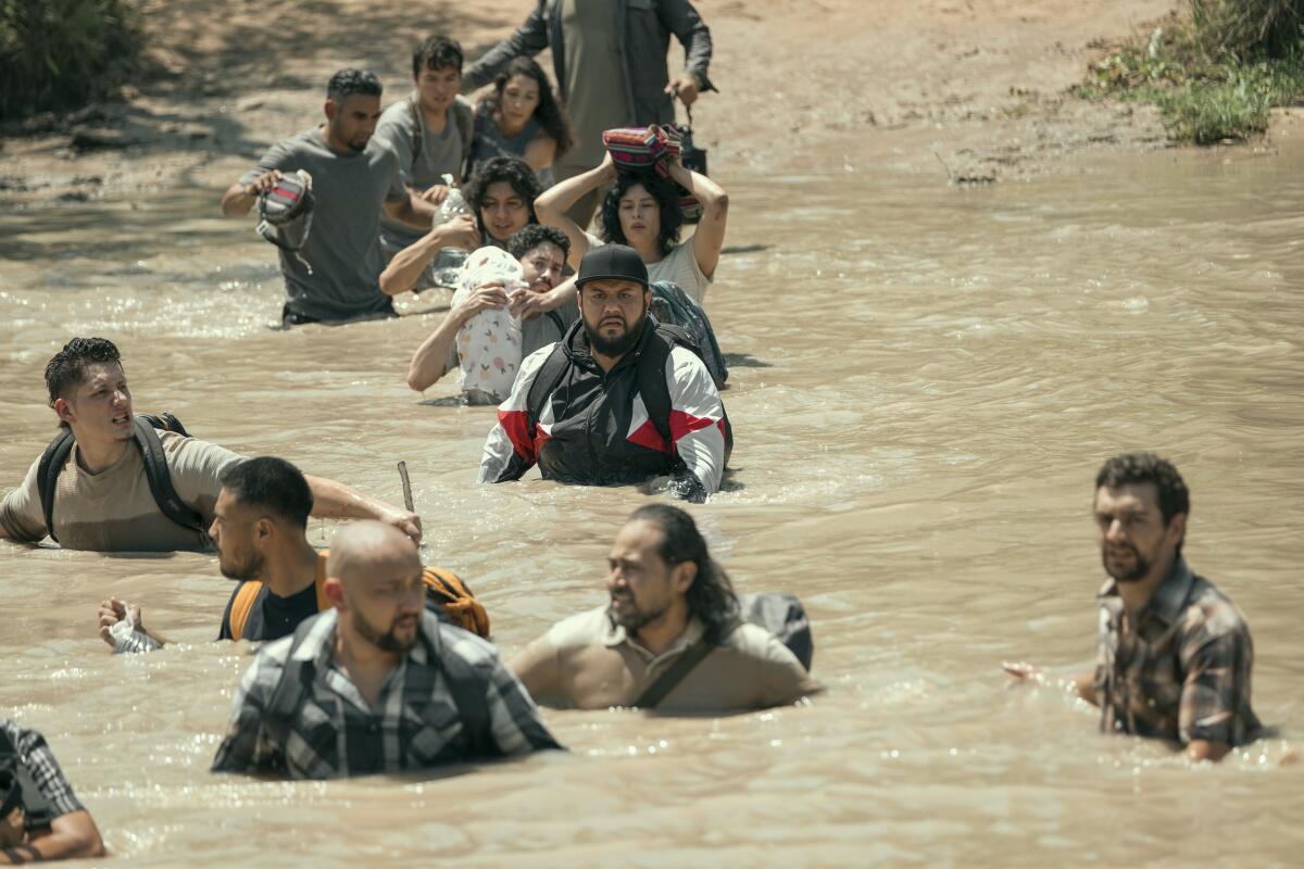 A man in a black ball cap and white, red and black jacket crosses a river with several men, women and children.