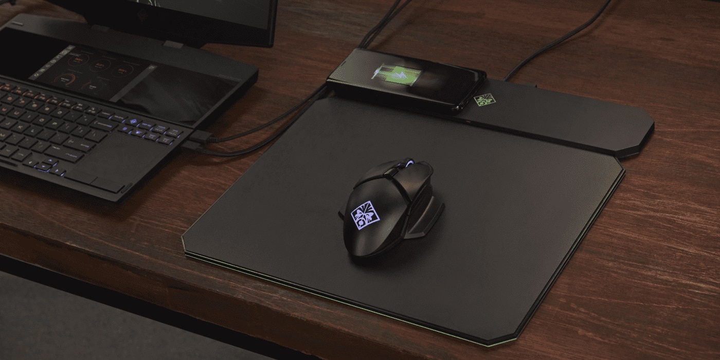 A gaming mouse with illuminated logo sits on an RGB-edged mousepad next to a smartphone wirelessly charging and a backlit gaming laptop on a wooden desk.