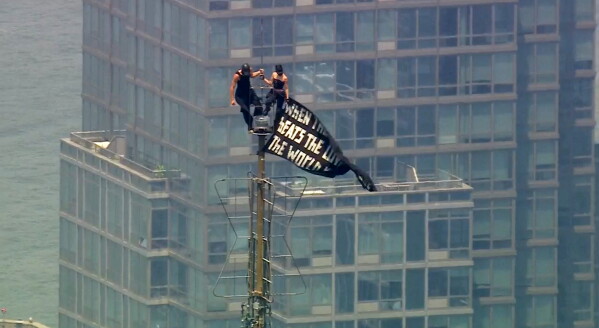 Two people stand on the tip of the antenna of the Empire State Building in New York while holding a banner on Wednesday, July 1, 2026. (WABC-TV via AP)