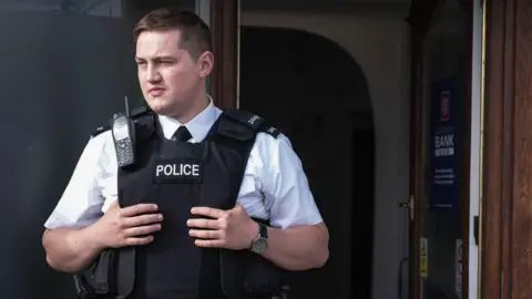 BBC/Long Story TV Finnian Garbutt, a man with dark hair, wearing a white shirt with black tie and a black support vest with the words police on it. He is wearing a black watch on his left wrist and there is a brown wooden door in the background.