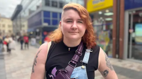 Freya Rocks has half of their head shaved, and the other half is long red hair. They wear a black sleeveless top and cross body bag,