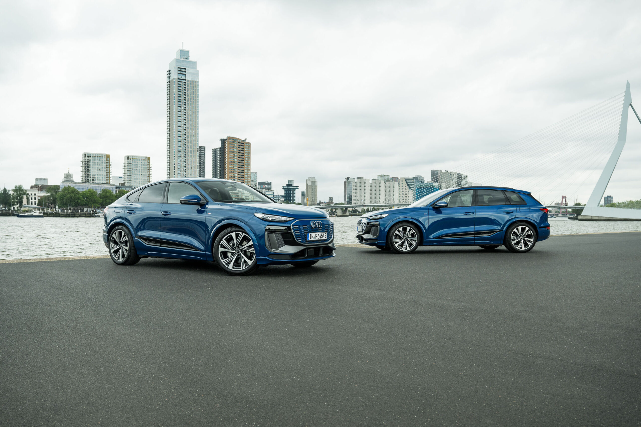 Audi Q6 e-tron Lifecycle Experience - Static shot: two Audi Q6 e-tron (Ascari blue), SUV and Sportback, shown in side and three-quarter views in an urban waterfront setting with a modern skyline and bridge in the background.