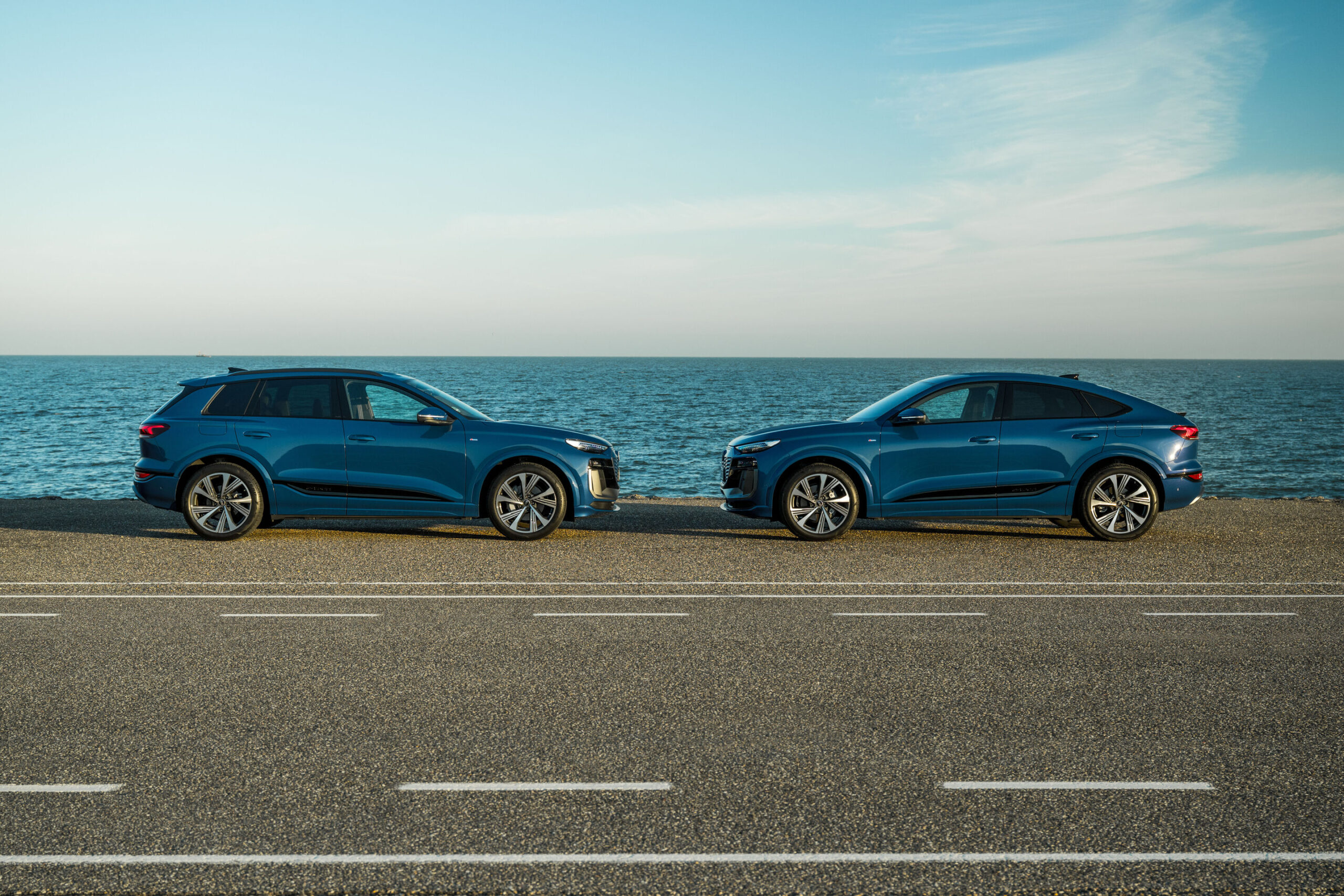 Audi Q6 e-tron Lifecycle Experience - Static shot: two Audi Q6 e-tron (Ascari blue), SUV and Sportback, shown in side profile facing each other on a coastal road with the open sea in the background.