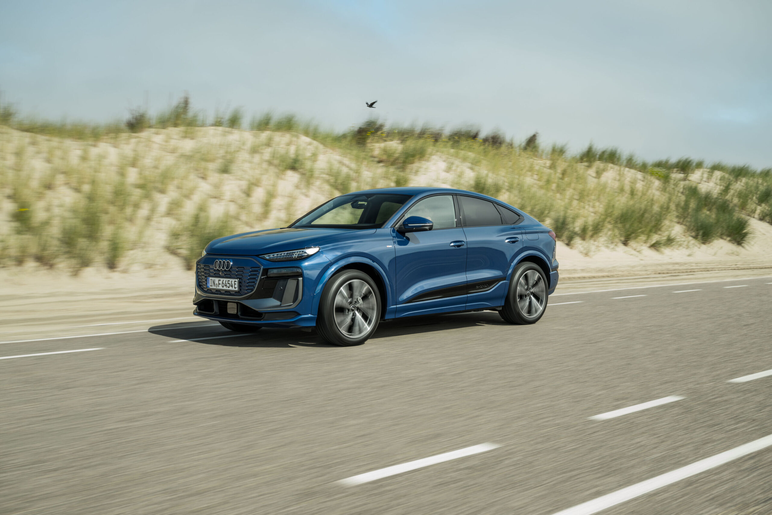 Audi Q6 e-tron Lifecycle Experience - Dynamic shot: Audi Q6 e-tron Sportback (Ascari blue), front three-quarter view in motion on a road alongside dune landscapes.