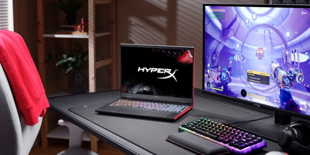 A HyperX gaming laptop with RGB keyboard sits on a dark desk beside a RGB mechanical keyboard and a monitor displaying a first-person shooter game, in a moody gaming setup.