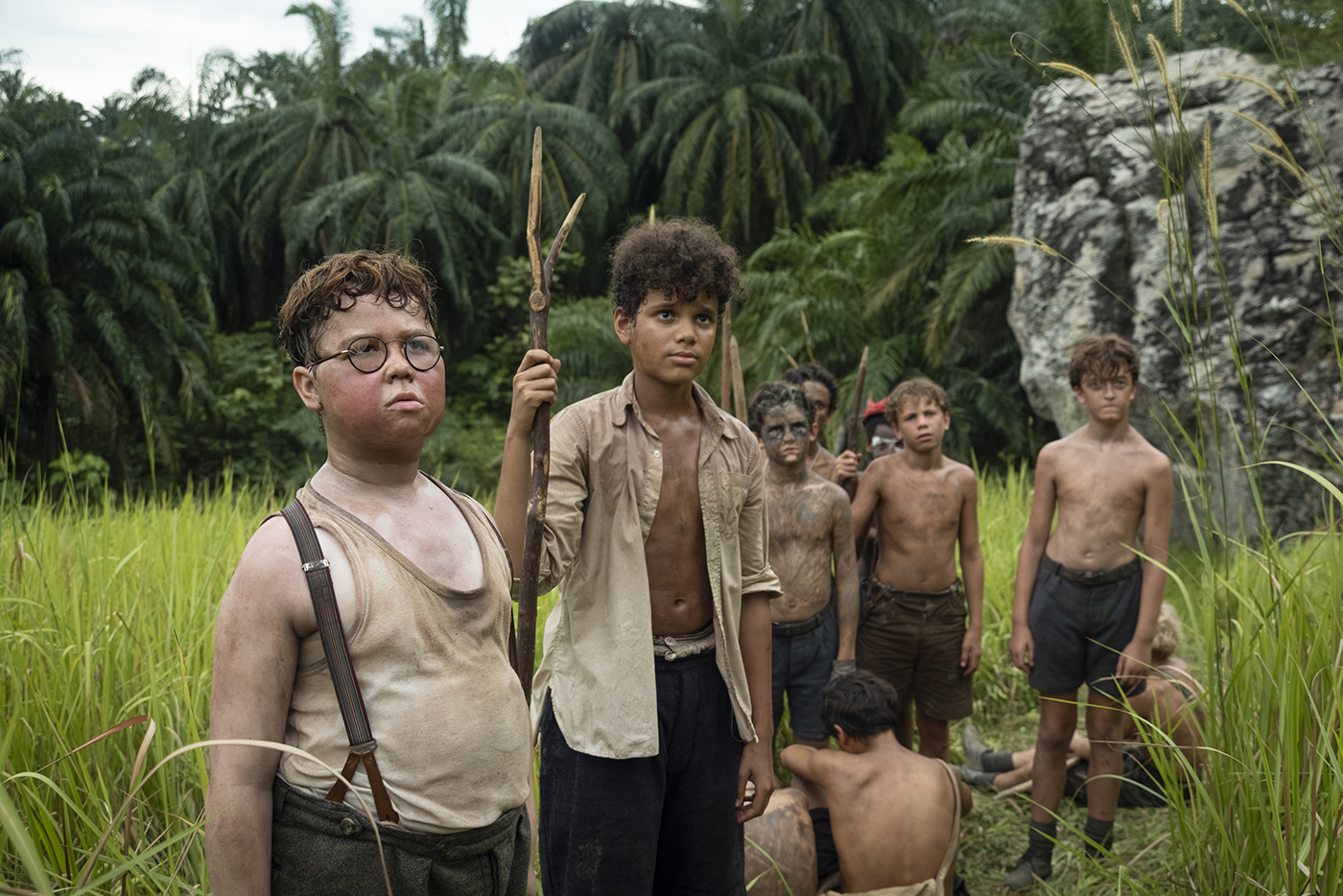 &Quot;A Group Of Dirty, Shirtless Boys Stand In Tall Grass With Jungle Foliage And A Rock Behind Them. Some Hold Makeshift Spears, Appearing Serious And Alert, As If Stranded Or Surviving In The Wild.&Quot;