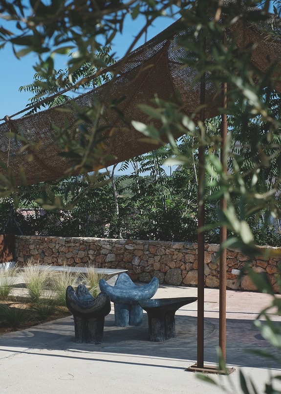Outdoor seating area with artistic stone chairs and table under a shade canopy, surrounded by trees and stone walls.