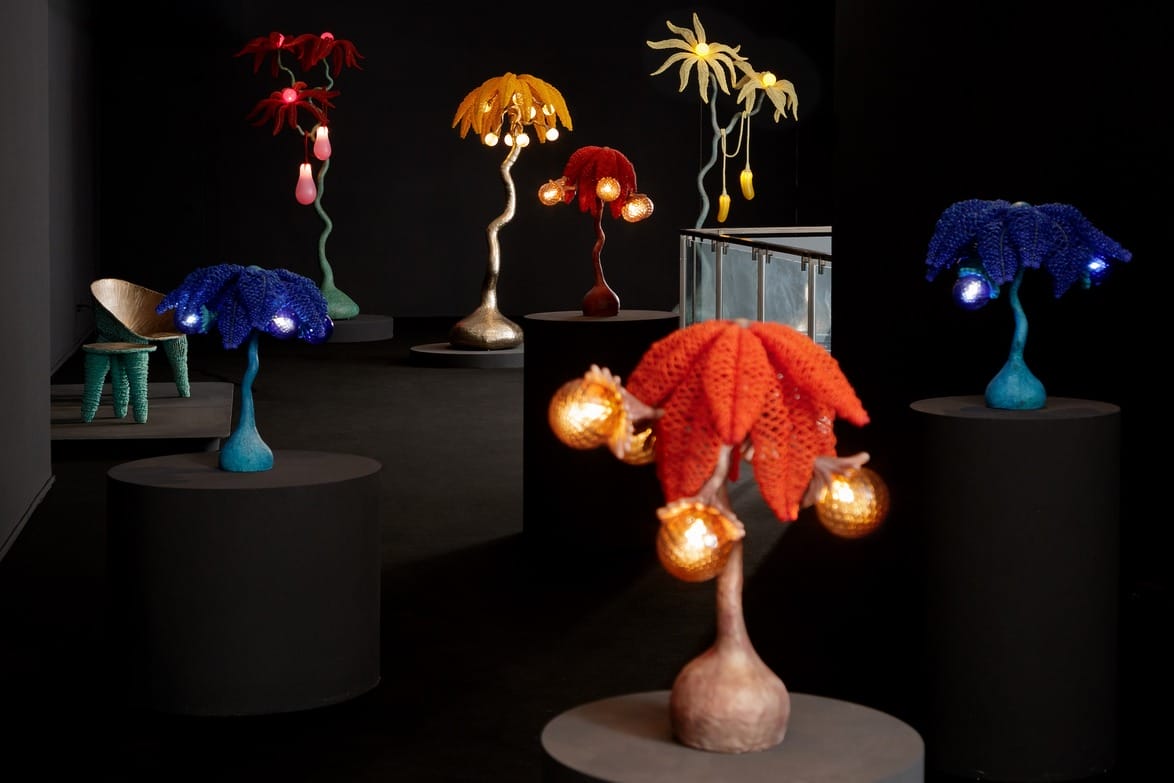 Colorful, abstract lamp sculptures with bulbous lights on display in a dark exhibition space with black cylindrical stands.