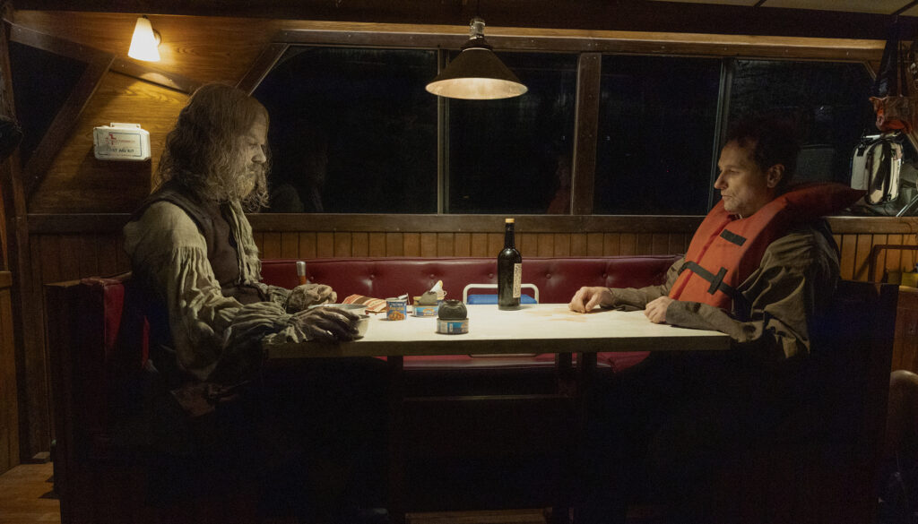 Widow's Bay Mayor Loftis sits opposite the town's tattered, 300-year-old unburied founder inside a dimly lit boat, capturing the show's strange mix of supernatural dread and deadpan humor.