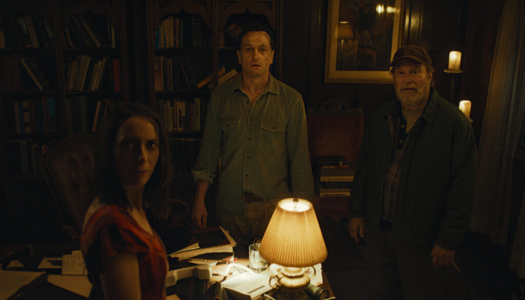 Still from Widow's Bay showing Patricia, Mayor Tom Loftis, and local Wyk standing in a dark, atmospheric wood-paneled library. They are looking directly forward with tense, wary expressions, dramatically lit by a single warm desk lamp in the foreground.