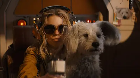 DC Studios A scene from Supergirl shows the title character sitting in a chair, wearing large, vintage over-ear headphones and large sunglasses in a darkened room. Her adorable white terrier sits on her lap looking alert.