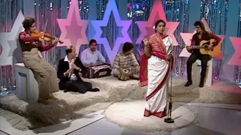 A woman in a white and red sari singing into a microphone while musicians are sat behind her playing instruments. The stage is decorated with large colourful stars and shiny curtains.