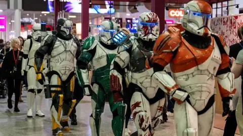 Getty Images Cosplayers dressed as Clonetroopers and Stormtroopers from the Star Wars films walking across the floor of a convention. They are wearing white armour, mixed with other colours, such as orange or green.
