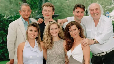 Getty Seven people arranged in two rows in a garden. Four people stand in the back row and three sit or stand in front. The group is posed closely together, with several people resting their hands on the shoulders of others. Most of the group are looking directly towards the camera.