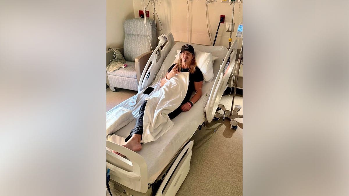 Bret Michaels in a hospital bed in an all-black outfit including a black hat.