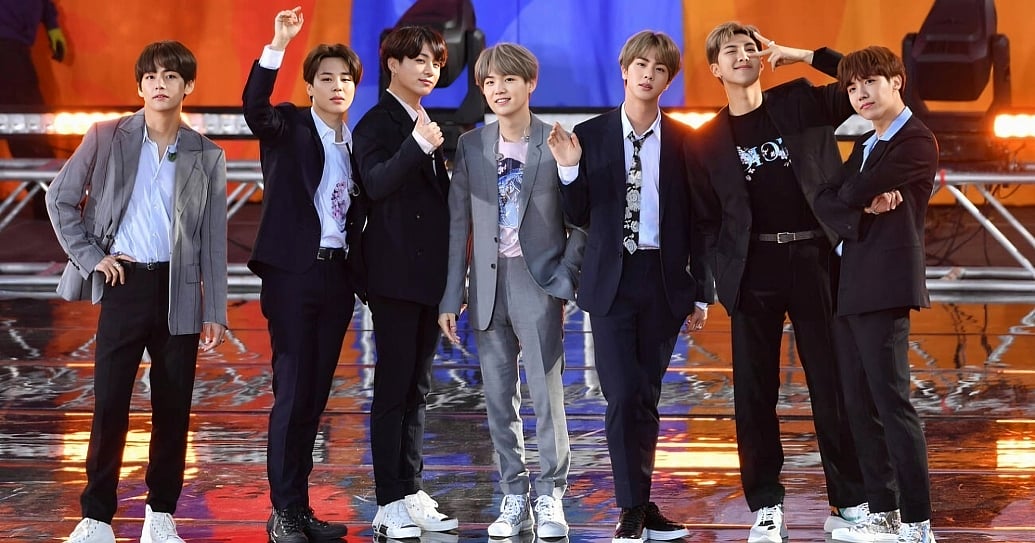 A full-body group photograph of the seven members of BTS standing side-by-side on a glossy, reflective stage. They are dressed in a coordinated mix of smart-casual suits, featuring black and grey blazers layered over button-down shirts, graphic t-shirts, and a patterned tie, completed with casual sneakers. The members smile and pose dynamically for the camera, with gestures including a hand waved high, a thumbs-up, and a peace sign. The background shows stage infrastructure illuminated by warm orange and bright blue lighting.
(L-R) Kim Tae-hyung, Park Ji-min, Jungkook, Suga, Kim Seok-jin, RM and J-Hope of BTS perform on "Good Morning America's Summer Concert Series" from Rumsey Playfield in Central Park on May 15, 2019 in New York City.