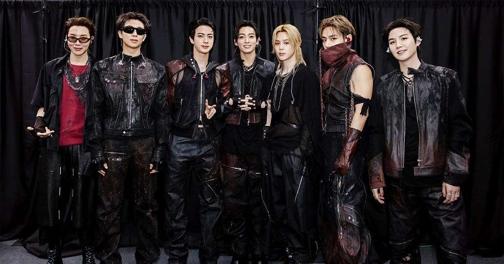 260612 BTS WORLD TOUR 'ARIRANG' IN BUSAN. A group photograph of seven young men standing side-by-side in a line against a backdrop of heavily pleated black curtains. They are wearing coordinated, edgy streetwear and tactical performance outfits in black, dark brown, and deep red tones, heavily detailed with straps, zippers, and layered silver chains. The two men on the far left are wearing black sunglasses. The group poses confidently under bright, direct lighting, with some individuals smiling and others sporting neutral expressions. BTS members RM (알엠, Kim Namjoon/김남준), Jin (진, Kim Seokjin/김석진), SUGA (슈가, Min Yoongi/민윤기), j-hope (제이홉, Jung Hoseok/정호석), Jimin (지민, Park Jimin/박지민), V (뷔, Kim Taehyung/김태형), and Jung Kook (정국, Jeon Jungkook/전정국).