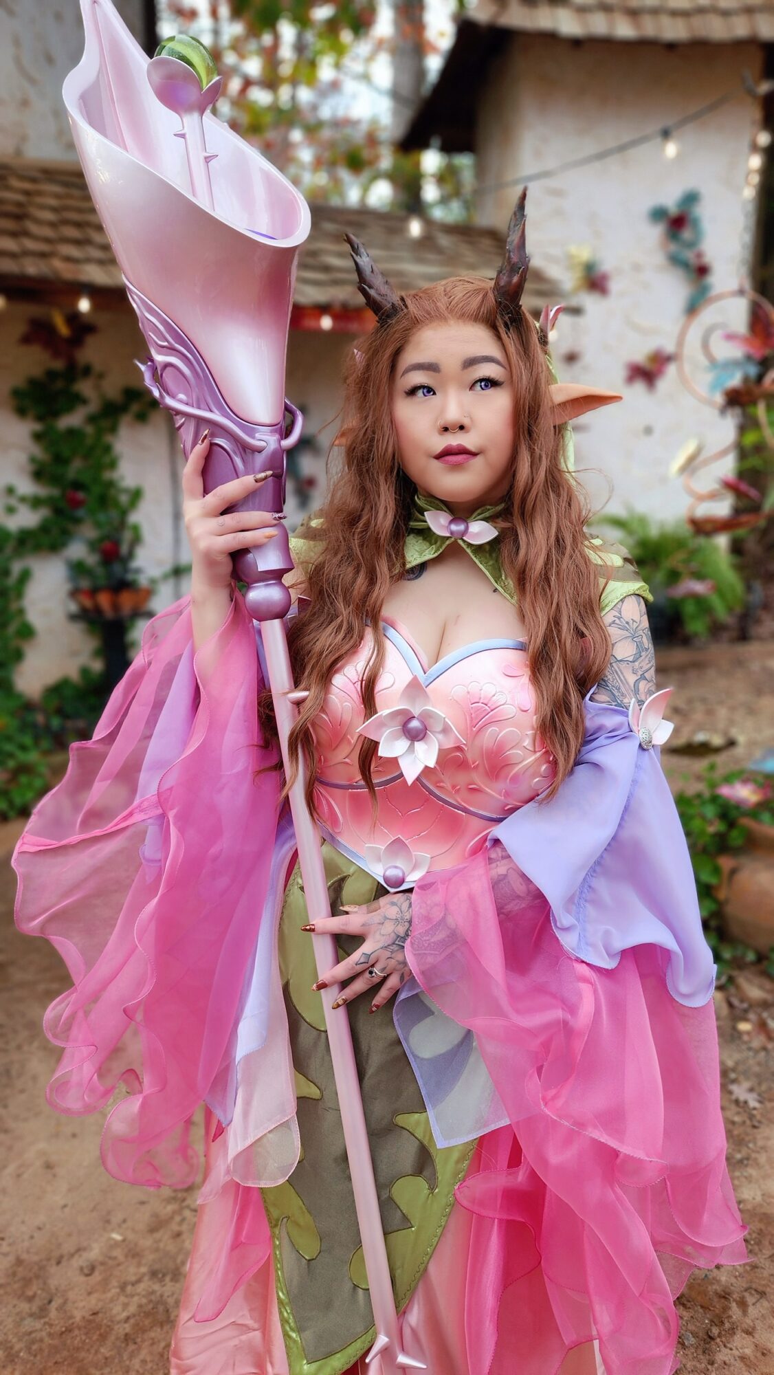 Young woman in colorful fantasy costume holding a large pink and purple staff, outdoors with plants and a building in background.