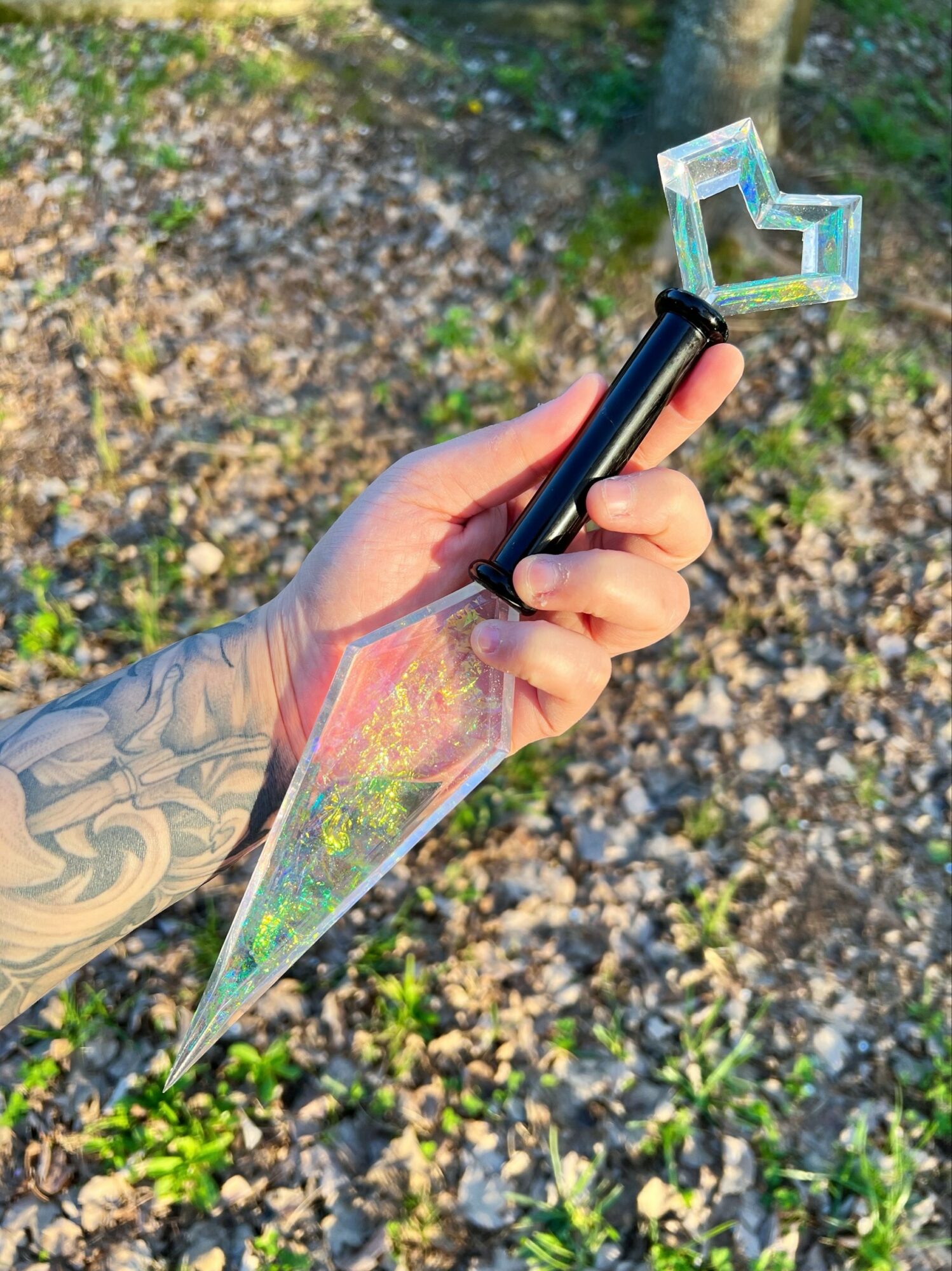 Person with a tattooed hand holding a transparent, iridescent dagger-shaped object outdoors, with a tree and ground covered in leaves and grass.