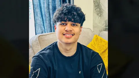 Anil Kumar Bharath A young man sat on a beige sofa with a yellow cushion beside them. He is wearing a dark sweatshirt with white geometric designs, with a blue curtain and leaf‑patterned wallpaper behind them.