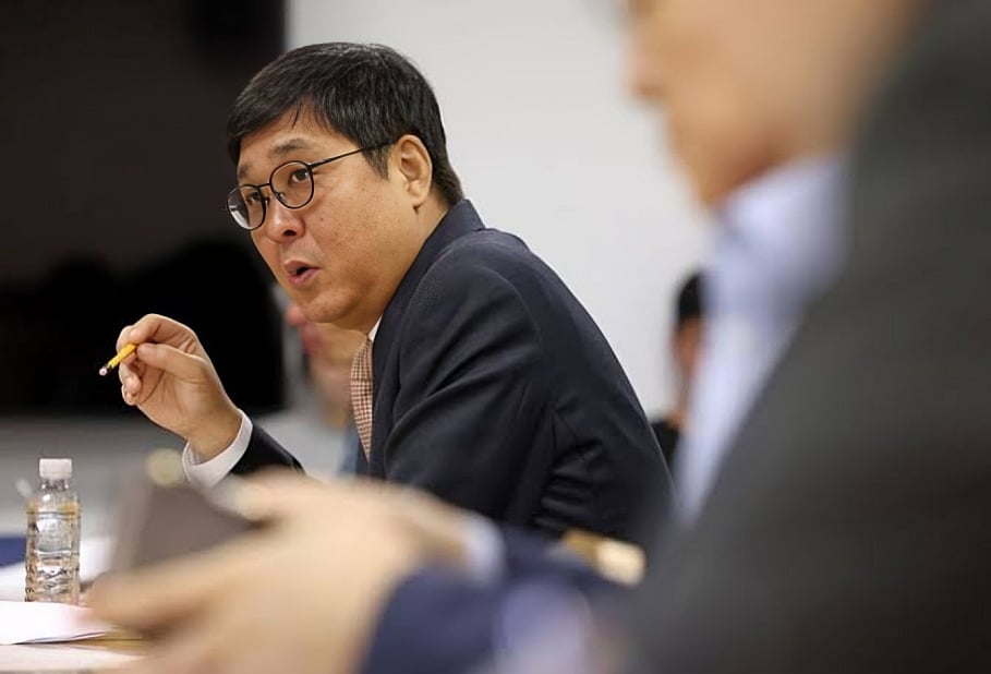 Medium shot of a South Korean Culture Minister Choi Hwi-young, wearing round glasses, a navy suit jacket, and a patterned tie, gesturing with a pencil in his hand as he speaks during a professional meeting. A water bottle sits on the table beside him, with blurred figures of other attendees visible in the foreground and background.