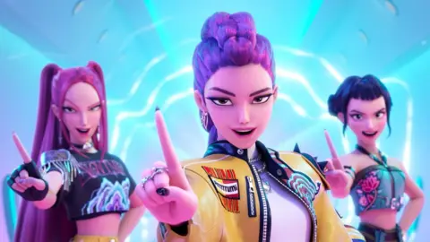 Netflix The animated characters from KPop Demon Hunters, as they appear in the film. They are dancing, mid-performance, holding their hands out towards the viewer, with brightly-coloured clothing and hair and under bright blue lighting.