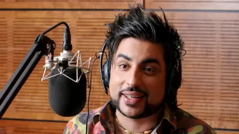 A male radio broadcaster wearing headphones speaking into a studio microphone mounted on an adjustable arm. He is seated in front of wooden acoustic panels.