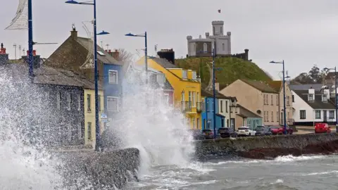 Getty Images An image of the seafront at Donaghadee, colourful houses can be seen along the promenade while waves break against the sea wall and spray up onto the road. It is an overcast day.