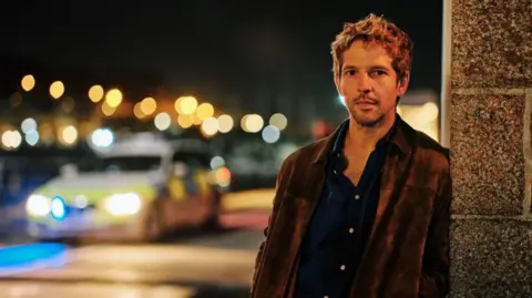 UKTV Actor Damien Molony in character as Jim Bergerac. He is leaning on a brick wall with a police car with its lights on blurred in the background. It is night time.