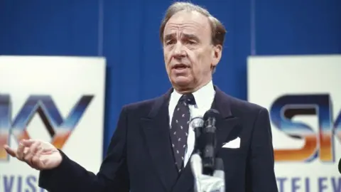 Getty Images Rupert Murdoch pictured at the launch of Sky Television in 1989. He is dressed in a black suit and black tie with white flecks on it. The Sky logo is seen behind him.