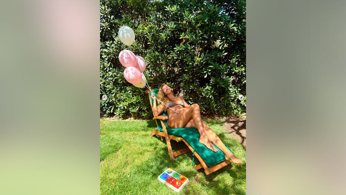 Gisele Bundchen on a green lounge chair holding balloons and smiling.