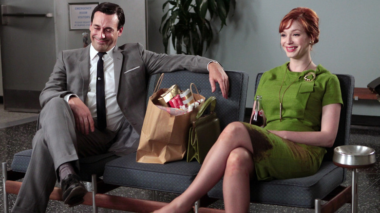 Jon Hamm as Don Draper and Christina Hendricks as Joan, sitting next to each other with a bag of gifts, smiling, in Mad Men