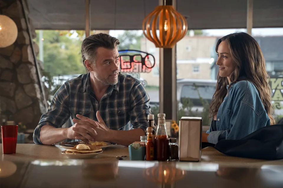 Josh Duhamel and Minka Kelly sit together at a diner counter in a scene from Ransom Canyon. Duhamel, wearing a blue plaid button-down shirt, looks toward Kelly with his hands folded beside a plate of pancakes. Kelly, dressed in a loose blue overshirt over a dark top, smiles back at him. Condiment bottles, a napkin dispenser and a coffee cup sit on the counter between them, while large windows and warm pendant lighting create a cozy, small-town diner atmosphere.
