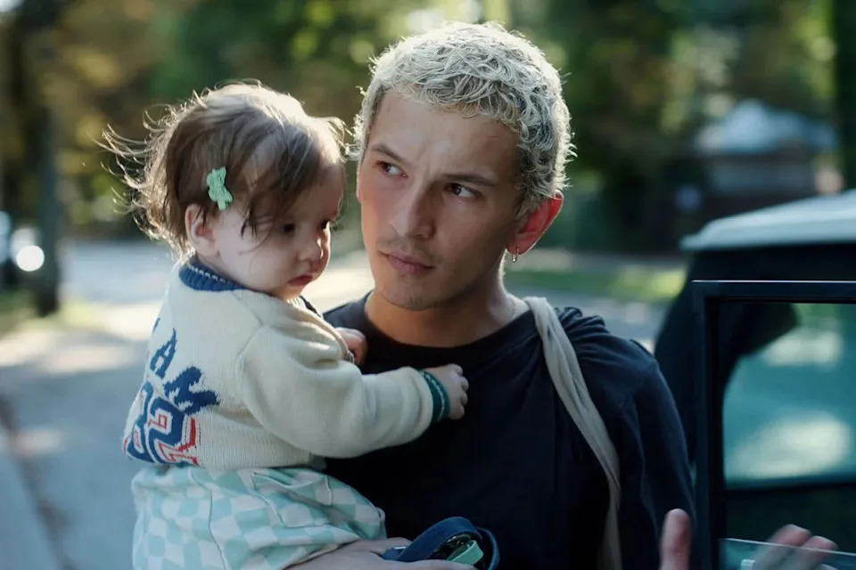 Filip Raczyński in a scene from "Proud." Young man with short bleached hair carries a toddler girl while standing next to a car.
