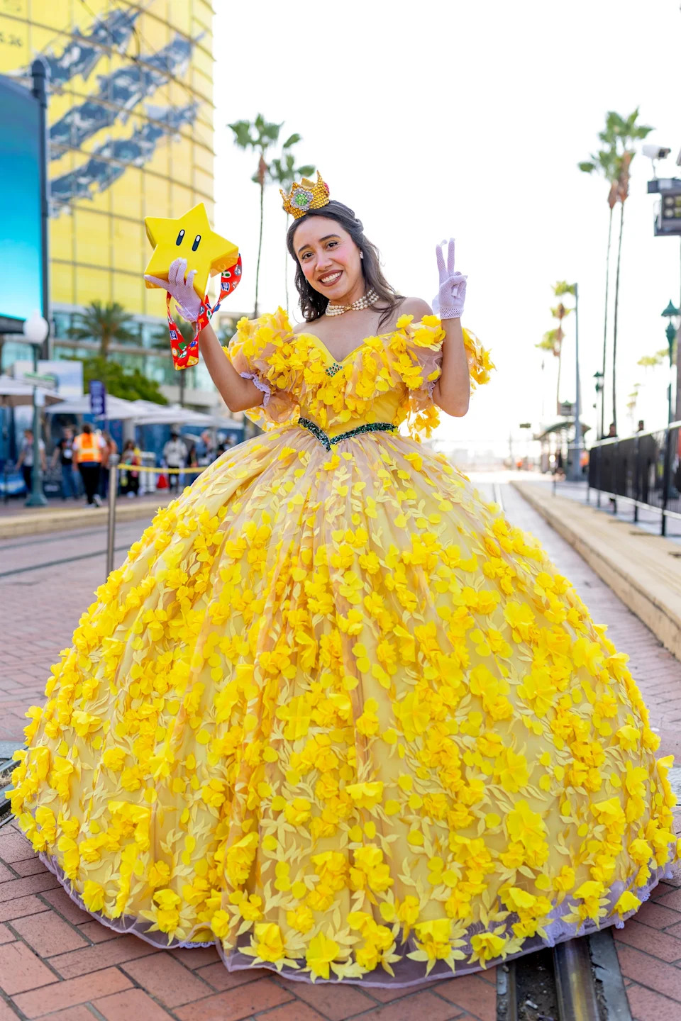 Person in a voluminous yellow floral dress, with crown and star, posing with peace sign outdoors