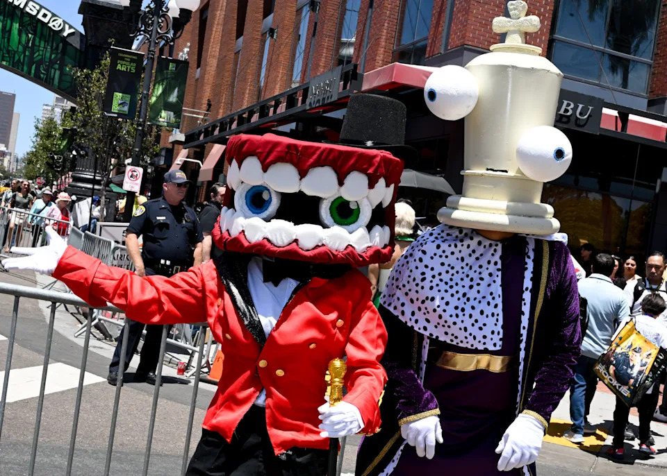 Two people in cartoonish costumes with oversized heads at an outdoor event. One wears a red suit, the other a purple robe