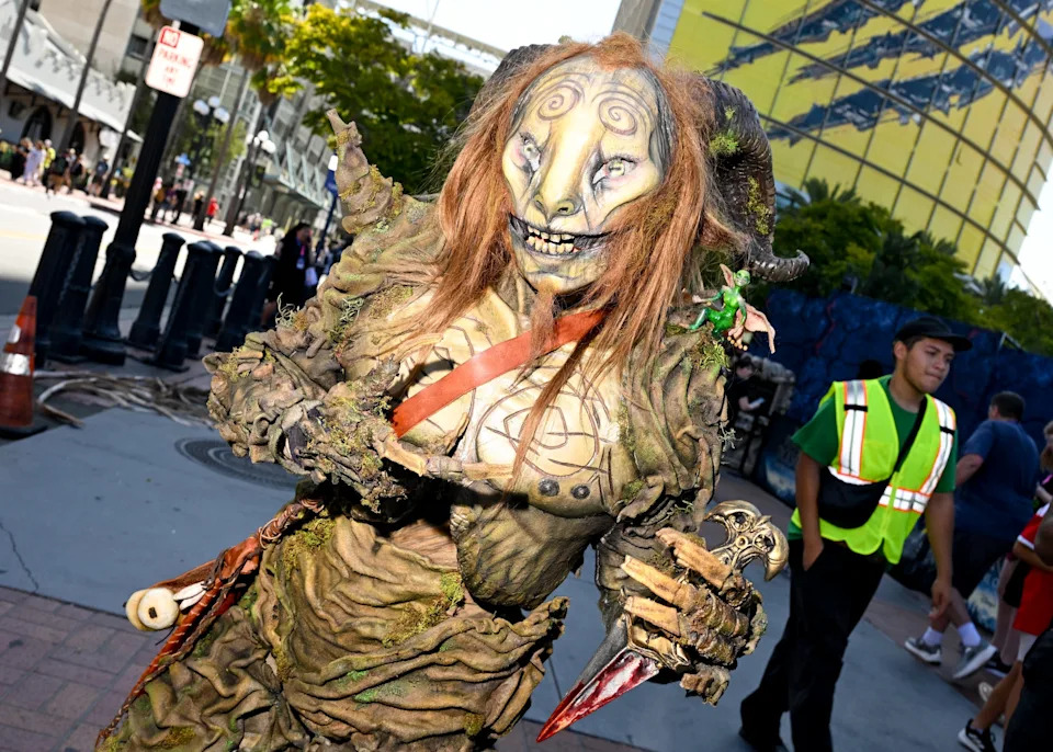 Person in intricate fantasy costume with a horned mask, detailed leafy textures, and a menacing expression, on a busy street
